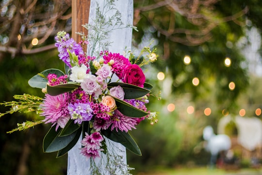 A vibrant floral arrangement featuring a variety of colorful flowers, including pink, purple, and white blooms, is set against a lush green natural backdrop. The bouquet is tied to a wooden frame with a white cloth, and soft, warm bokeh lights create a dreamy atmosphere in the background.