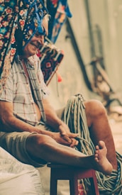 An elderly man with a content expression is seated on a stool, surrounded by colorful textiles and ropes. He wears a short-sleeved plaid shirt and shorts, and is barefoot. The setting appears rustic and relaxed, with a focus on leisure.