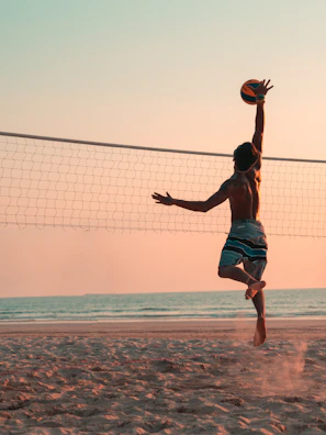 A player leaping high for a powerful spike during a beach volleyball match at sunset.