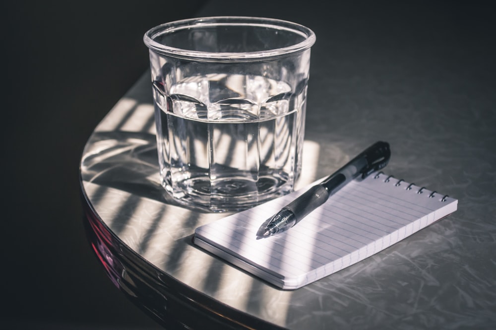 clear drinking glass beside pen and paper