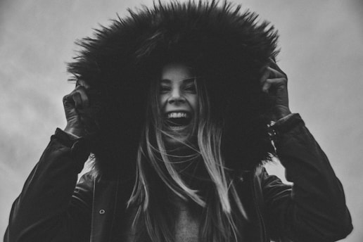 A person is laughing joyfully, holding the hood of a fur-lined coat. The image is in black and white, highlighting the textures and contrasts.
