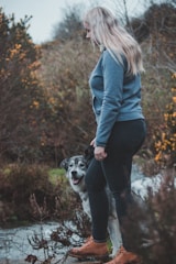 A woman wearing a blue sweater and dark jeans is standing next to a large dog in a natural setting with shrubs and trees. The dog is looking towards the camera with a cheerful expression. The woman is wearing brown boots and appears to be near a small stream or path.