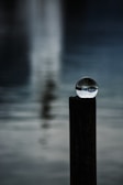 A clear glass sphere is delicately balanced on top of a dark wooden post. The background features a blurred, serene body of water with subtle reflections.