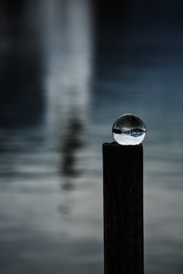 A clear glass sphere is delicately balanced on top of a dark wooden post. The background features a blurred, serene body of water with subtle reflections.