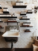 A vintage-style washroom with a weathered, metal-framed sink. Multiple shelves with neatly arranged bottles line the tiled wall, giving a rustic and organized aesthetic. The background consists of white subway tiles and exposed patches, suggesting an industrial or retro interior design.