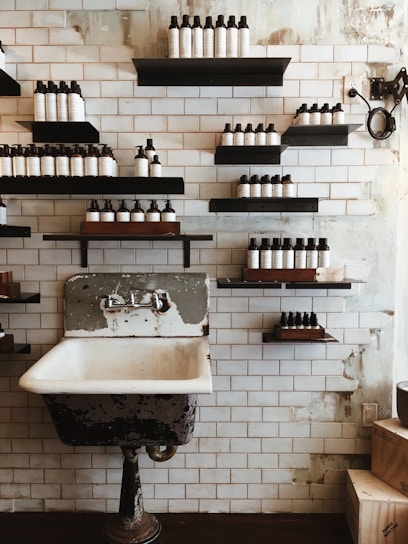 A vintage-style washroom with a weathered, metal-framed sink. Multiple shelves with neatly arranged bottles line the tiled wall, giving a rustic and organized aesthetic. The background consists of white subway tiles and exposed patches, suggesting an industrial or retro interior design.