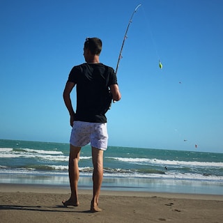 A person stands on a sandy beach holding a fishing rod, facing the ocean. The water features gentle waves under a clear blue sky. The individual appears relaxed, wearing a black shirt and white shorts.