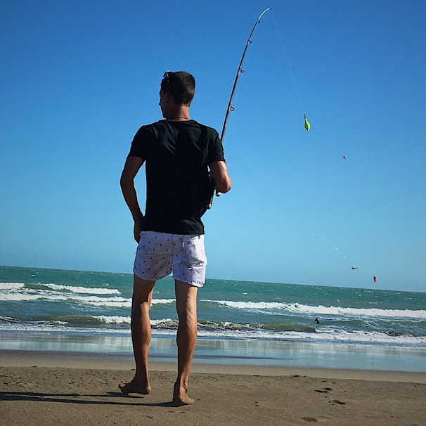 A person stands on a sandy beach holding a fishing rod, facing the ocean. The water features gentle waves under a clear blue sky. The individual appears relaxed, wearing a black shirt and white shorts.