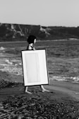 grayscale photo of woman walking on seashore white holding rectangular wooden photo frame