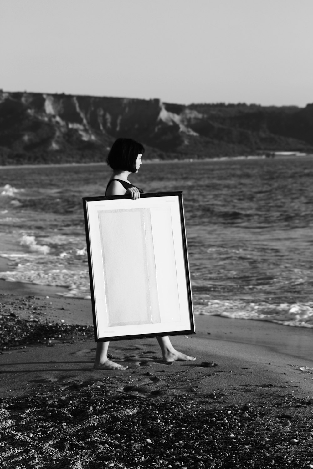Foto em tons de cinza da mulher que caminha na costa branca segurando moldura retangular de madeira