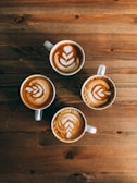 Four cups of coffee with latte art patterns arranged neatly on a wooden table. Each cup features a similar leaf-like motif created with steamed milk, showcasing skilled barista work.