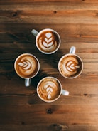 Four cups of coffee with latte art patterns arranged neatly on a wooden table. Each cup features a similar leaf-like motif created with steamed milk, showcasing skilled barista work.
