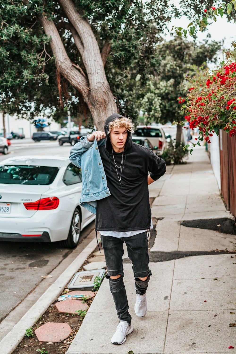 man wearing blue denim jacket while walking on the street near green tree