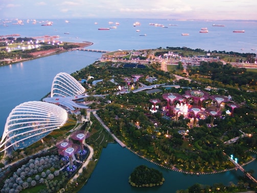 Aerial view of a landscaped garden with futuristic structures and domes beside a large body of water filled with ships. The area is lush with greenery and features several illuminated towers and pathways.