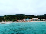 Along a sandy beach, several colorful buildings and huts line the shore, backed by lush green hills under a cloudy sky. The turquoise sea in the foreground contains a boat, displaying a flag.