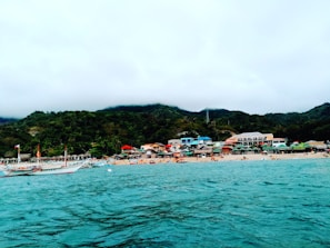 Along a sandy beach, several colorful buildings and huts line the shore, backed by lush green hills under a cloudy sky. The turquoise sea in the foreground contains a boat, displaying a flag.