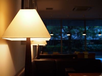 A warmly lit lamp stands prominently in the foreground, casting a soft glow. In the background, more lamps are lined up, creating a cozy ambiance in what seems to be a dimly lit indoor setting, possibly a restaurant or cafe. Outside the large windows, the sky is dark, and the interior appears calm and inviting.