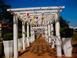 A pergola with white columns and overhead lattice adorned with colorful hanging lanterns extends over a tiled pathway. Large white planters with green shrubs line either side of the walkway. Shadows of the structure create patterns on the ground. The sky is clear and blue, while trees and buildings can be seen in the background.