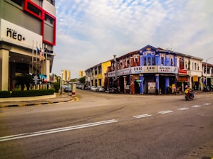 A street scene featuring an intersection with a hotel on the left and historic buildings on the right. The hotel has modern architectural elements, including red accents, while the buildings opposite have a vintage style with weathered facades. Flags are displayed in front of the hotel. A motorbike is visible on the right side, and there are other vehicles parked along the street.