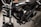 A close-up view of a black motorcycle, focusing on the engine and components. The chrome and metallic parts are polished, with the brand name prominently displayed on the side. The background appears to be a dark, textured surface.