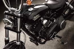 A close-up view of a black motorcycle, focusing on the engine and components. The chrome and metallic parts are polished, with the brand name prominently displayed on the side. The background appears to be a dark, textured surface.