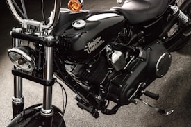 A close-up view of a black motorcycle, focusing on the engine and components. The chrome and metallic parts are polished, with the brand name prominently displayed on the side. The background appears to be a dark, textured surface.