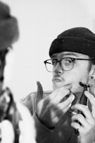 A person with glasses and a beanie is using an electric trimmer to shave facial hair while looking in a mirror. Their expression appears focused as they groom their chin, with one hand holding the trimmer and the other touching their face.