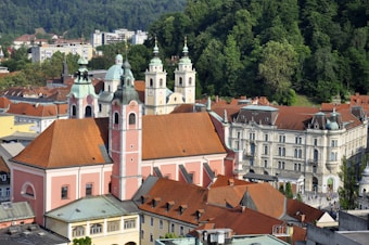 A picturesque view of a European city featuring a large, historic church with multiple towers and red-tiled roofs. The architecture is complemented by surrounding classical buildings and lush green hills in the background, indicating a blend of natural and urban landscapes.