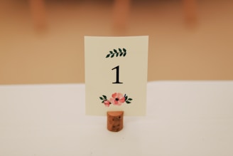 A table number sign with the number 1 displayed in black on a white card, adorned with floral designs in red and green. The card is held upright by a cork stand on a white tablecloth.