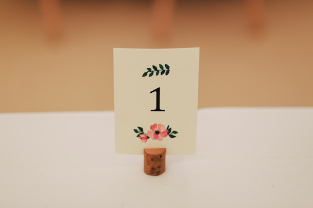 A table number sign with the number 1 displayed in black on a white card, adorned with floral designs in red and green. The card is held upright by a cork stand on a white tablecloth.