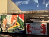 A mural featuring a charismatic figure raising a fist, next to a quote about going to prison before becoming President. The background includes flags and colors. Adjacent text addresses social and political issues, and there are images of two red figures facing each other against a white backdrop.