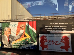 A mural featuring a charismatic figure raising a fist, next to a quote about going to prison before becoming President. The background includes flags and colors. Adjacent text addresses social and political issues, and there are images of two red figures facing each other against a white backdrop.
