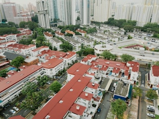 An aerial view of a residential area with a mix of low-rise buildings and high-rise apartment complexes. The buildings have distinctive red roofs, and there are well-organized streets and greenery scattered throughout the area. Numerous cars can be seen parked along the streets.