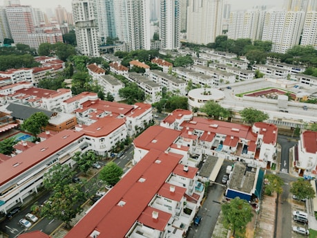 An aerial view of a residential area with a mix of low-rise buildings and high-rise apartment complexes. The buildings have distinctive red roofs, and there are well-organized streets and greenery scattered throughout the area. Numerous cars can be seen parked along the streets.