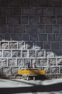 A stone wall composed of square gray bricks with a wooden sign leaning against it. The sign reads 'Day to day good home' in green text painted on a yellow background. The scene is partially shadowed, suggesting sunlight from an angled direction.