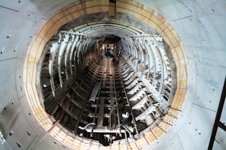 Inside view of a tunnel under construction, featuring curved walls lined with a structural framework. The perspective is straight down the tunnel, showing construction materials and scattered debris, with artificial lighting illuminating the scene.