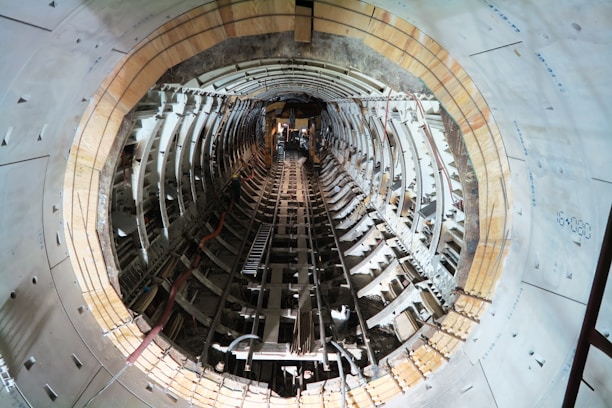 Inside view of a tunnel under construction, featuring curved walls lined with a structural framework. The perspective is straight down the tunnel, showing construction materials and scattered debris, with artificial lighting illuminating the scene.