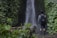A person stands facing a waterfall, surrounded by lush greenery. The individual is wearing a backpack and boots, suggesting a hiking context. The forest vegetation is dense, with various shades of green, and the waterfall creates a serene, natural setting.