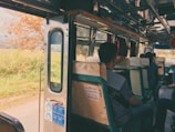 Inside view of a bus with passengers seated on old-fashioned green and white seats. The bus door is open, revealing a scenic view of a rural landscape with a tree and greenery outside. Sunlight filters in, creating a warm, nostalgic atmosphere.