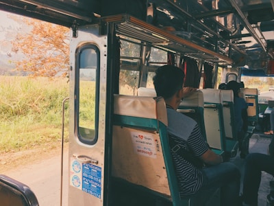 Inside view of a bus with passengers seated on old-fashioned green and white seats. The bus door is open, revealing a scenic view of a rural landscape with a tree and greenery outside. Sunlight filters in, creating a warm, nostalgic atmosphere.