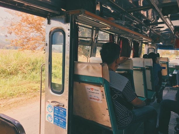 Inside view of a bus with passengers seated on old-fashioned green and white seats. The bus door is open, revealing a scenic view of a rural landscape with a tree and greenery outside. Sunlight filters in, creating a warm, nostalgic atmosphere.