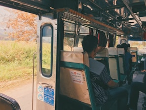 Inside view of a bus with passengers seated on old-fashioned green and white seats. The bus door is open, revealing a scenic view of a rural landscape with a tree and greenery outside. Sunlight filters in, creating a warm, nostalgic atmosphere.