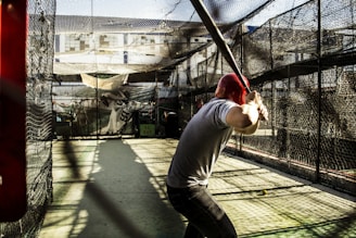 A person wearing a red helmet is poised in a batting stance with a baseball bat in a batting cage. The cage is enclosed with black netting and sunlight is casting shadows on the ground. The background shows some buildings and other equipment inside the cage.