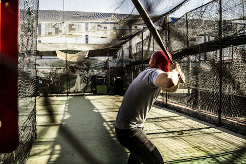 A person wearing a red helmet is poised in a batting stance with a baseball bat in a batting cage. The cage is enclosed with black netting and sunlight is casting shadows on the ground. The background shows some buildings and other equipment inside the cage.