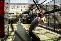 A person wearing a red helmet is poised in a batting stance with a baseball bat in a batting cage. The cage is enclosed with black netting and sunlight is casting shadows on the ground. The background shows some buildings and other equipment inside the cage.