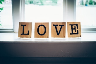 Wooden tiles with black letters spell out the word 'LOVE' while sitting on a windowsill. Each tile bears a small number in the corner, similar to Scrabble tiles. Soft natural light filters through the window, creating a cozy and serene atmosphere.