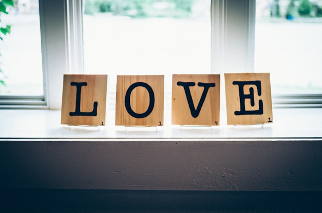 Wooden tiles with black letters spell out the word 'LOVE' while sitting on a windowsill. Each tile bears a small number in the corner, similar to Scrabble tiles. Soft natural light filters through the window, creating a cozy and serene atmosphere.
