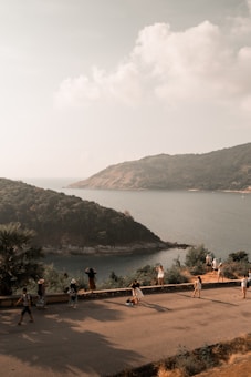 A scenic coastal landscape with a view of the ocean and a hill covered in greenery. Several people can be seen standing and walking along a road, some taking photos and enjoying the view. The sky is partly cloudy and the lighting suggests either early morning or late afternoon.