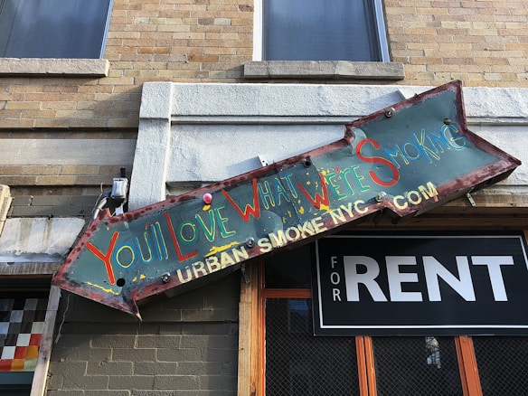 A weathered and colorful sign features the text 'You'll Love What We're Smoking' in vibrant letters. The sign has an arrow shape and is mounted on a building with a brick exterior. Below the sign, there is a 'FOR RENT' sign with a black background and white text.