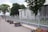An ornate iron fence painted in light gray runs along a paved sidewalk. Behind the fence is a historic building with arched green-trimmed windows. Flower pots with pink blooms are neatly lined up along the sidewalk, with verdant trees providing a lush backdrop.